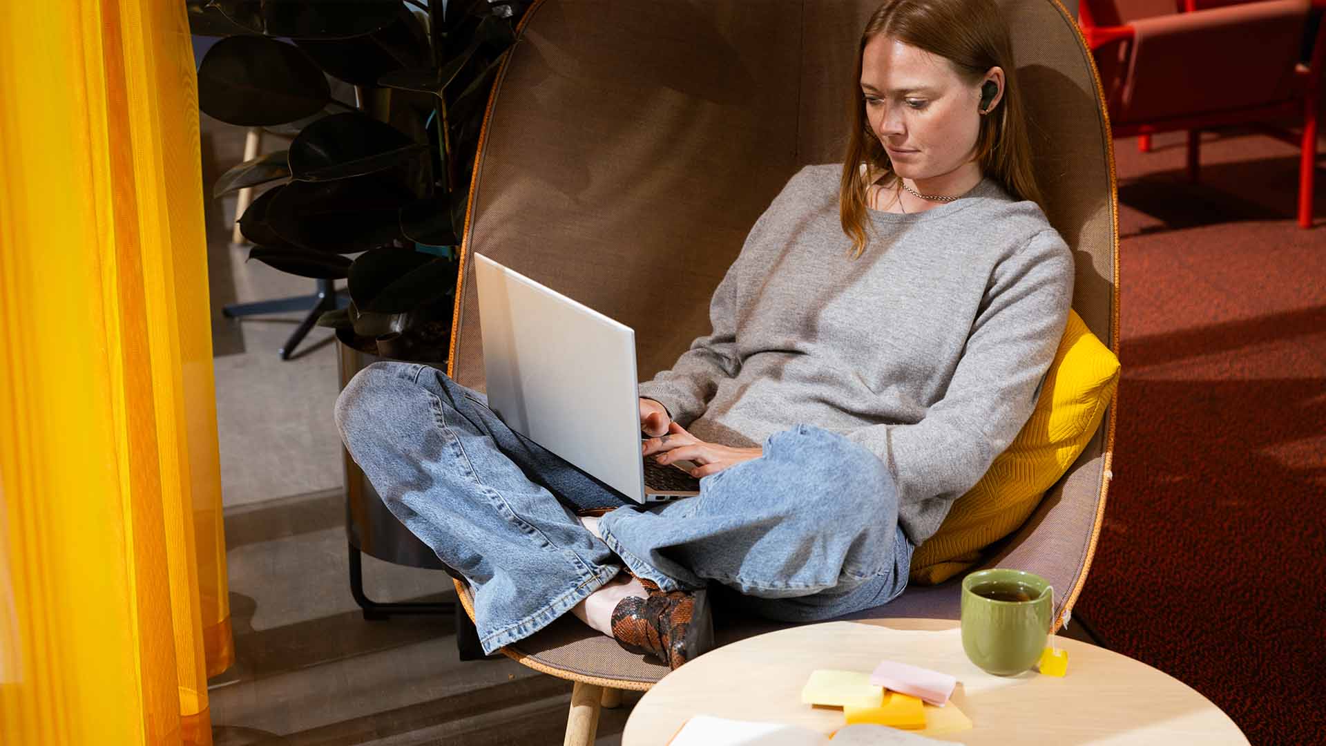 Kvinna i grå tröja sitter i stol med laptop i knät, gul kudde bakom ryggen. På bordet bredvid henne står en kopp te och det ligger post-it-lappar på bordet.