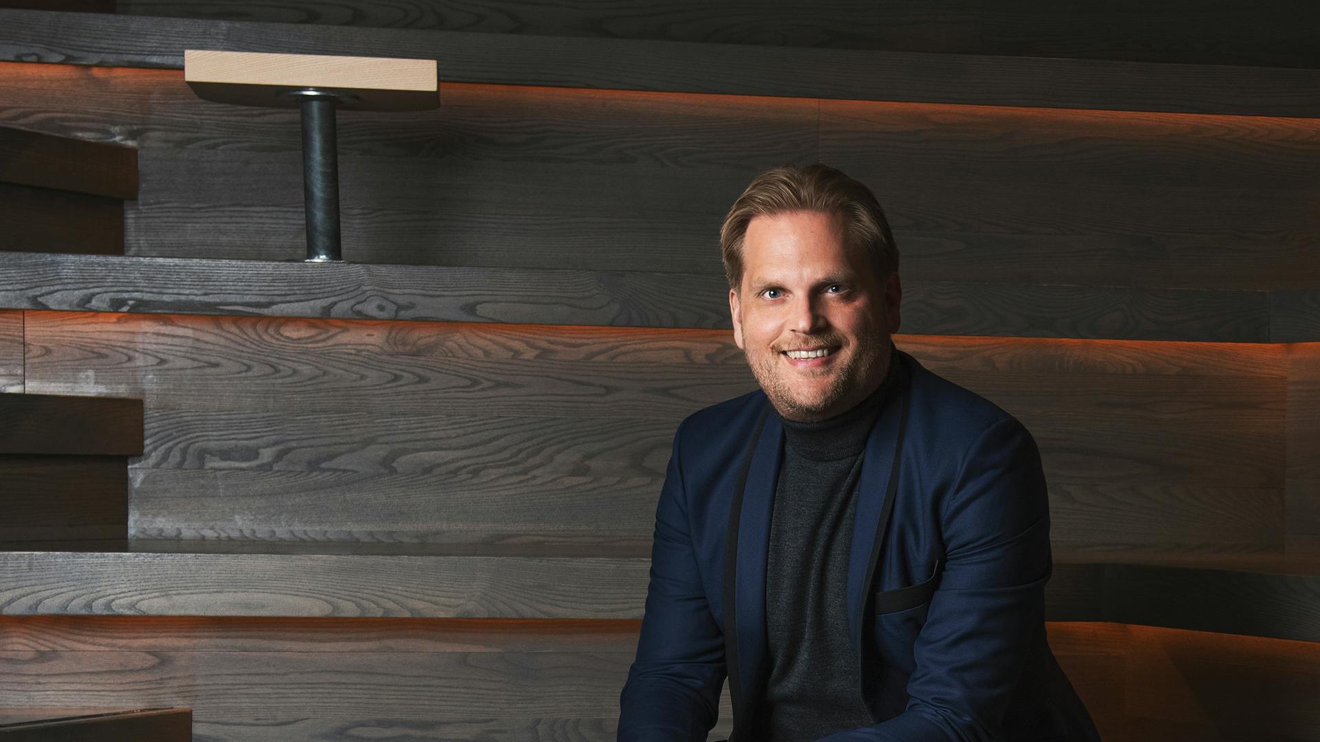 Christian Albinsson, iklädd kavaj och svarta kläder sitter med ett leende i en trappa med små bord. 