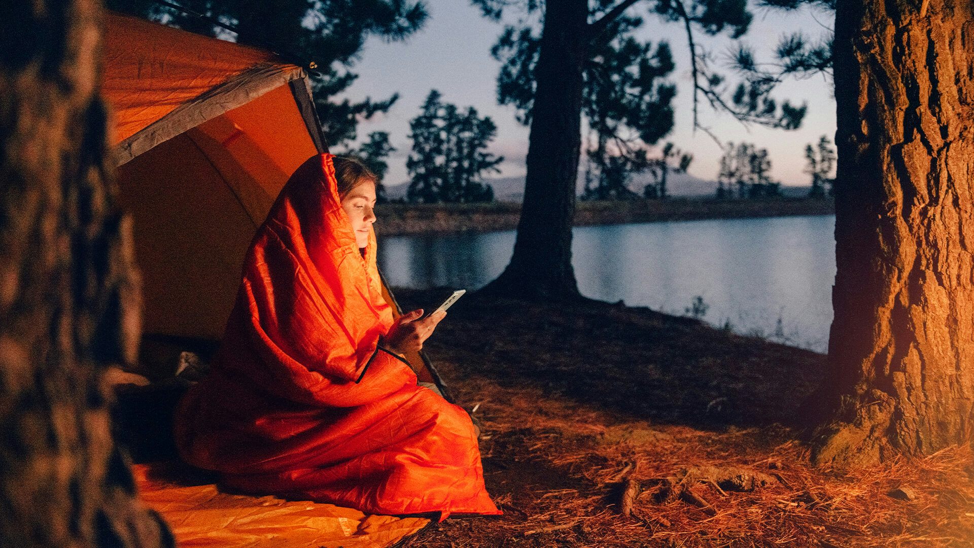 En kvinna sitter framför ett tält vid vattnet med en sovsäck runt sig. I handen håller hon en mobil som hon tittar på.