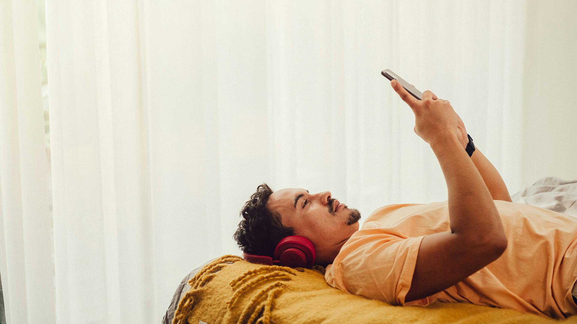 En man med hörlurar ligger på en säng och tittar på en mobil.
