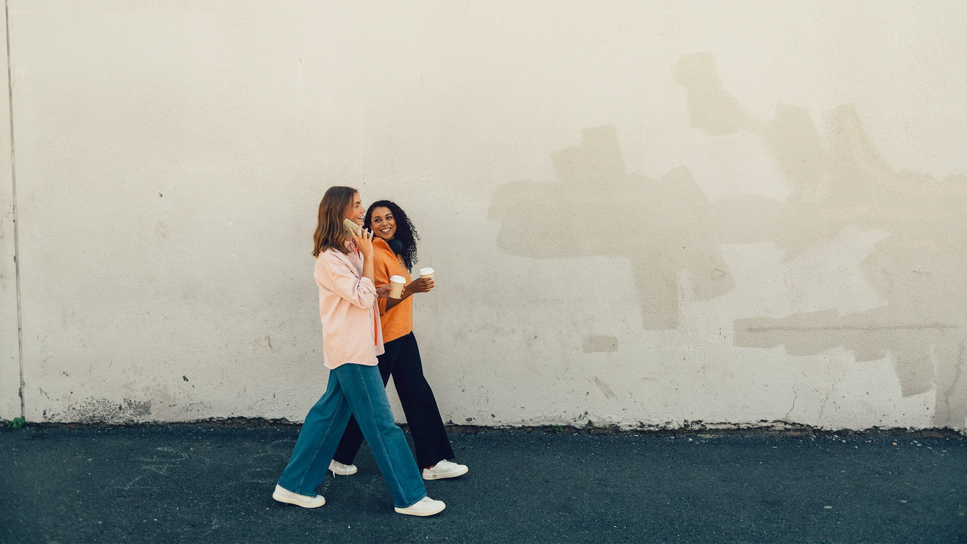 Två tjejer går med varsin kaffe i handen. Den ena pratar i telefon.