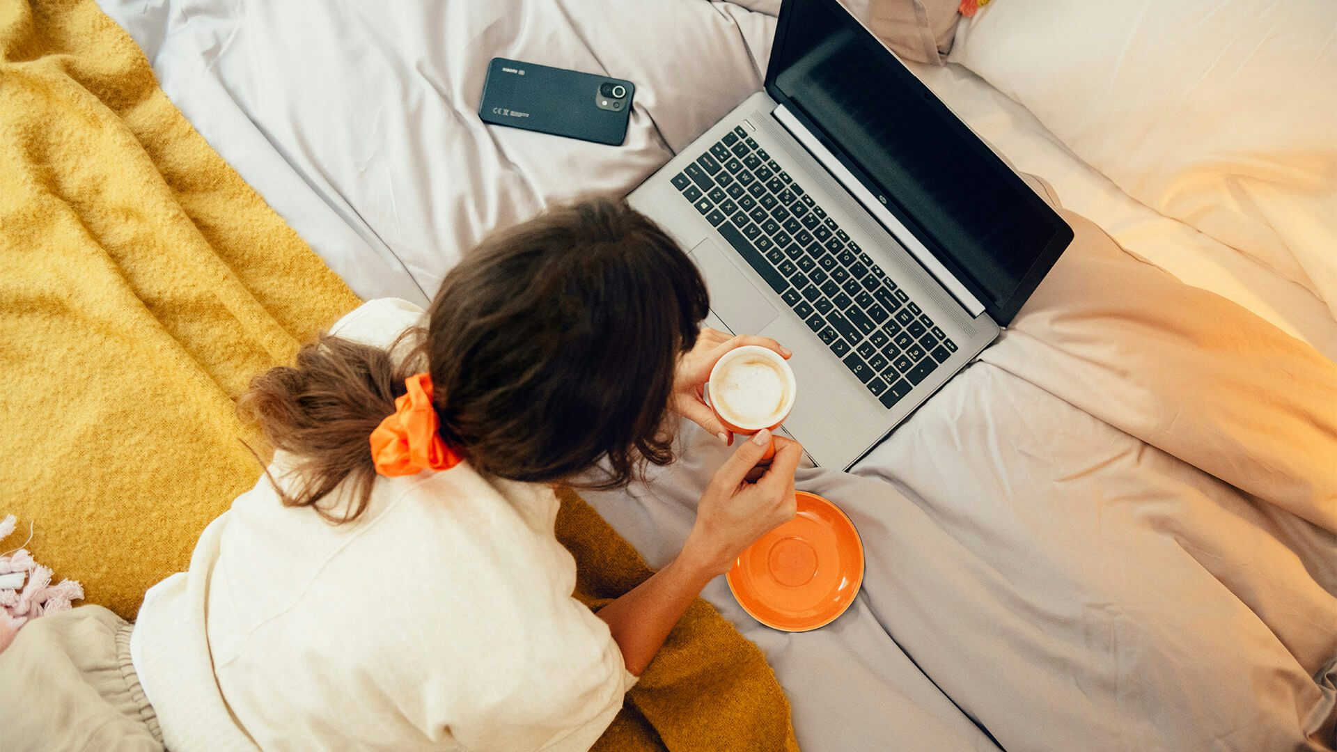 Kvinna dricker kaffe i sängen medan hon surfar på sin dator. Bredvid sig har hon sin mobil