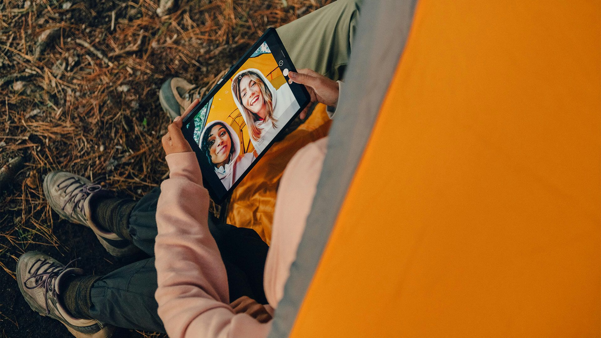 Två tjejer sitter i ett tält och tittar på sig själva i kameran på en surfplatta.