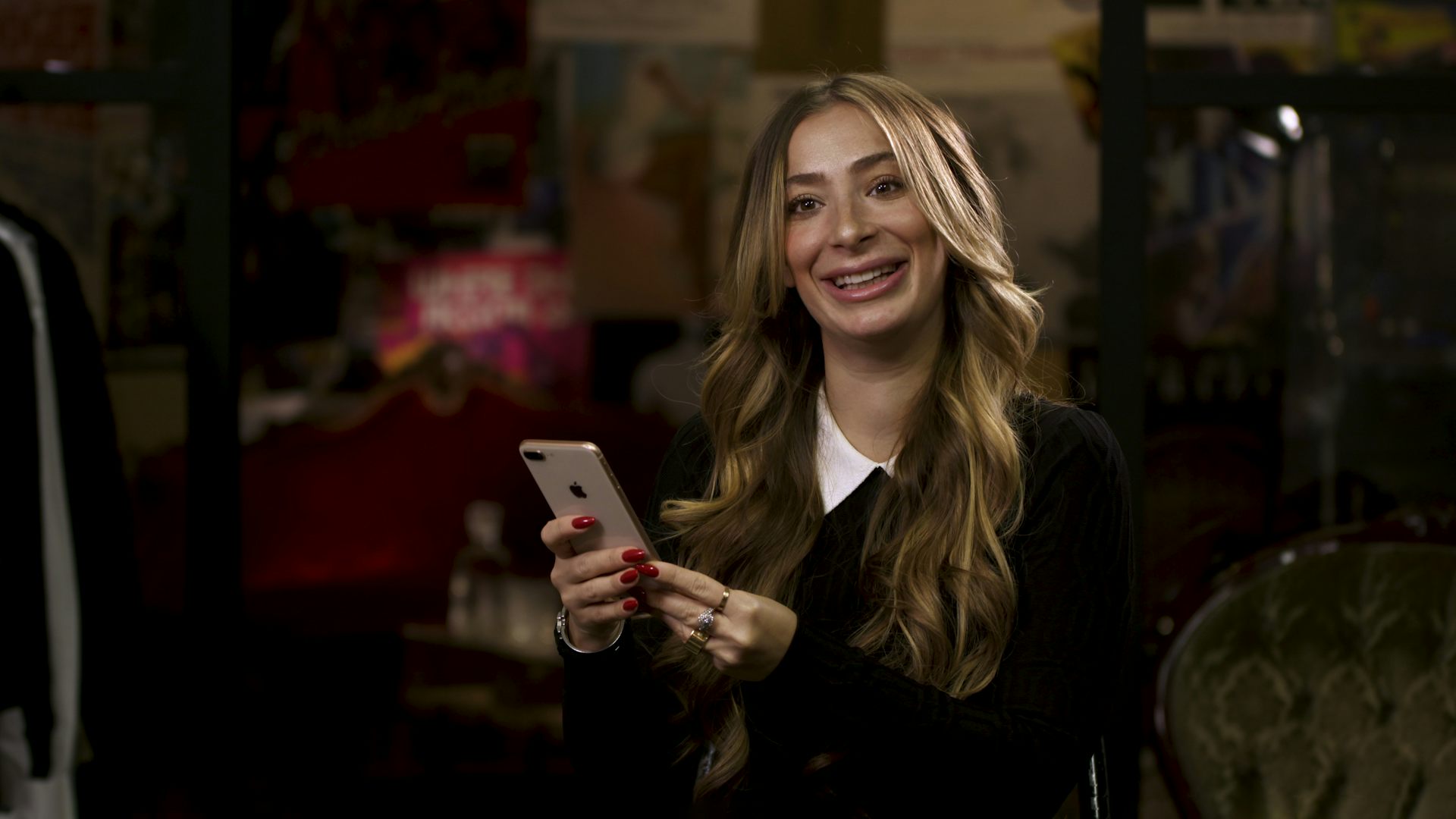 Kvinna i långt, lockigt, brunt hår iklädd svart pullover-tröja där en vit skjortkrage sticker ut håller i en smartphone. Hon tittar mot kameran och ler.