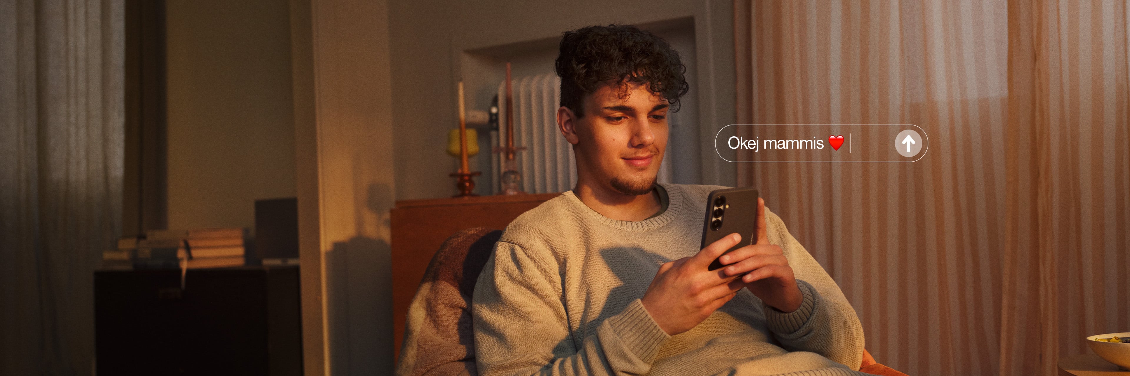 En kille i grå tröja och jeans sitter i en fåtölj och tittar i sin mobil.