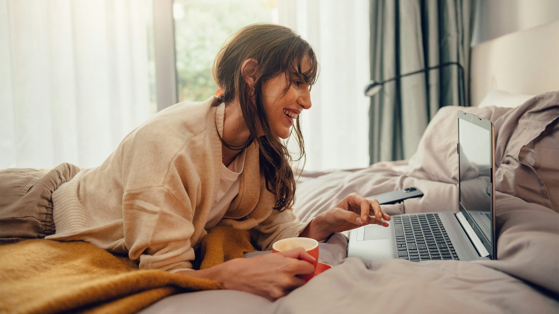 Kvinna ligger på en säng med en kaffekopp i handen och tittar på en dator.