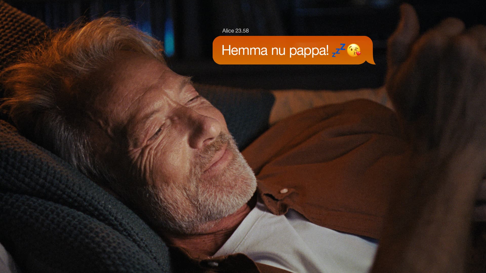 En äldre man ligger och sover med ett leende. Ett meddelande från "Alice" visas: "Hemma nu pappa! och två emojis.
