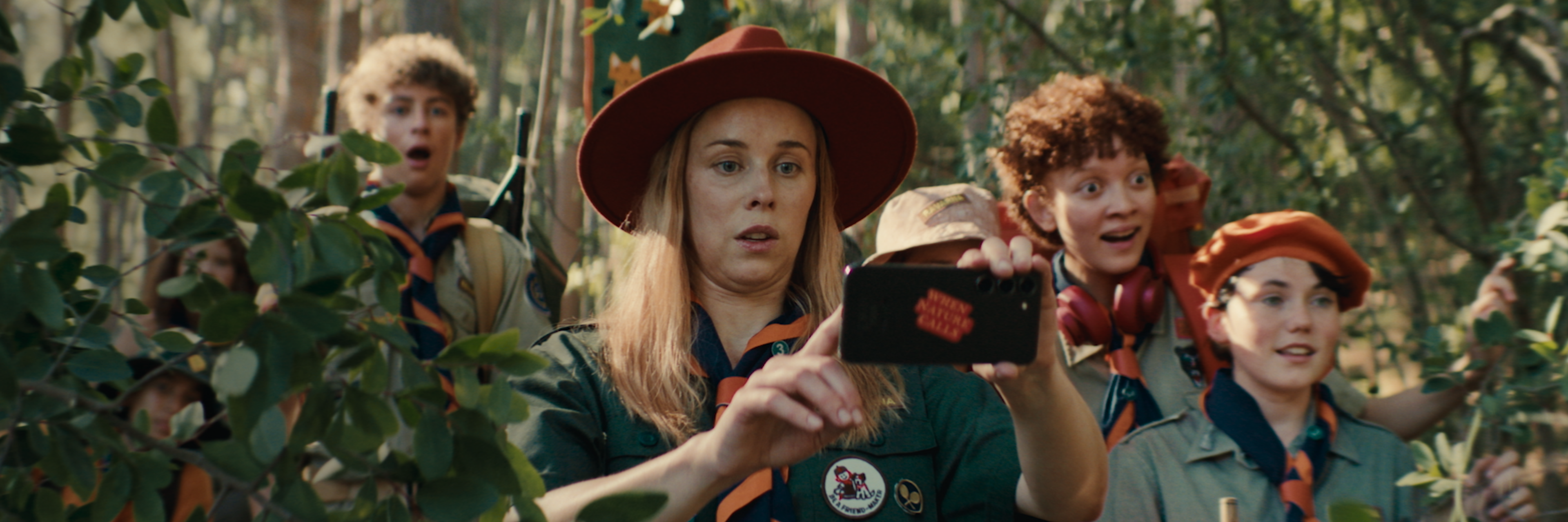 En scout står i skogen tillsammans med andra scouter och fotograferar med en svart mobil.