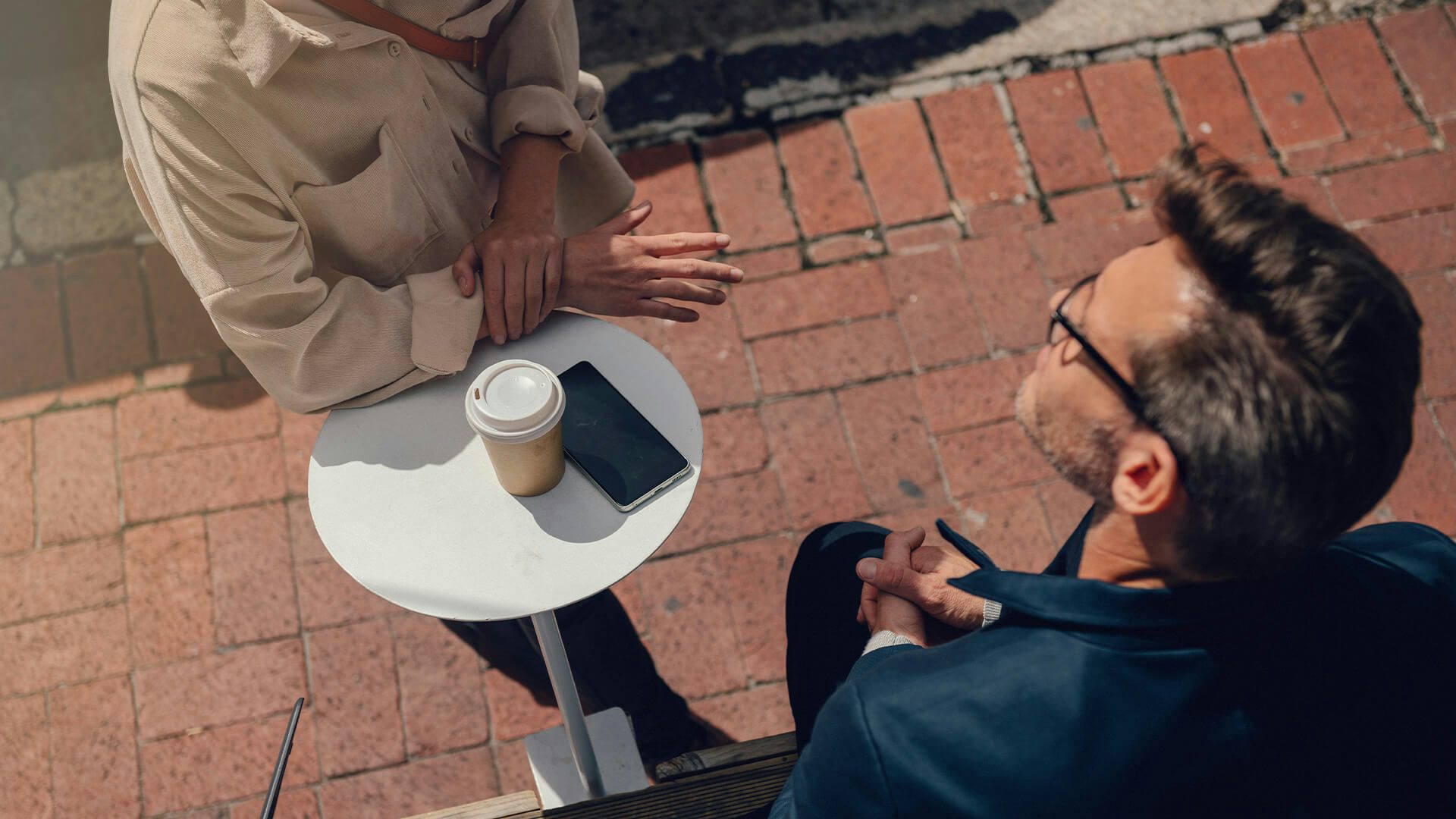 Två personer samtalar utomhus över en kaffe, en mobil ligger på ett vitt bord.