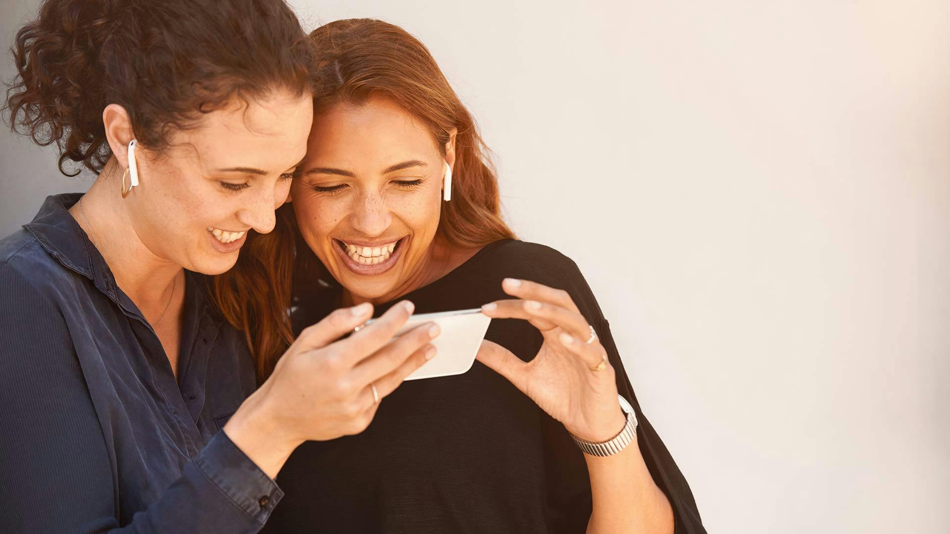 Två kvinnor med ett stort leende tittar på en mobil. De har airpods i öronen. 