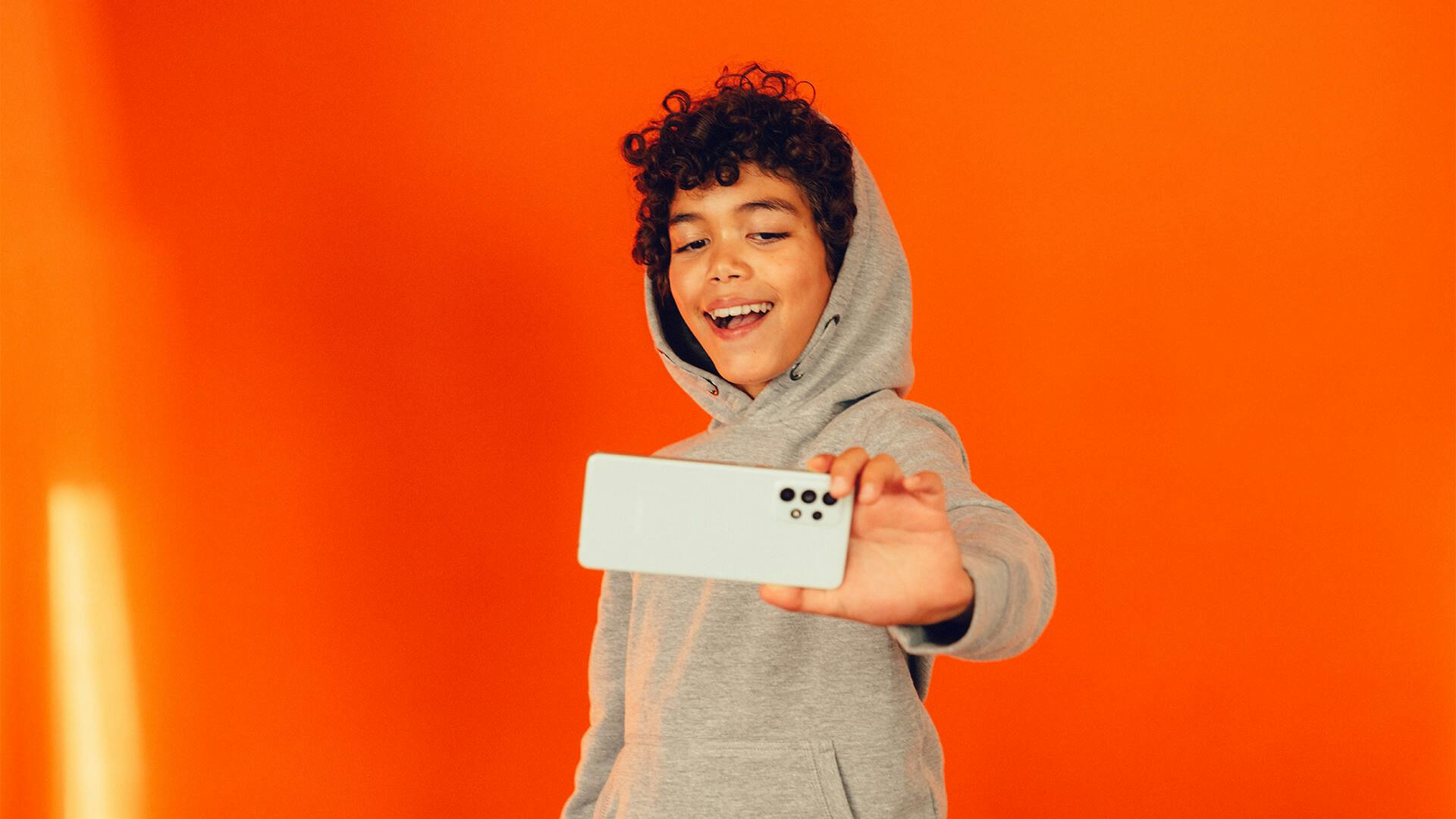 Barn ler mot mobilen som han håller framför sig. Bakgrunden är orange.