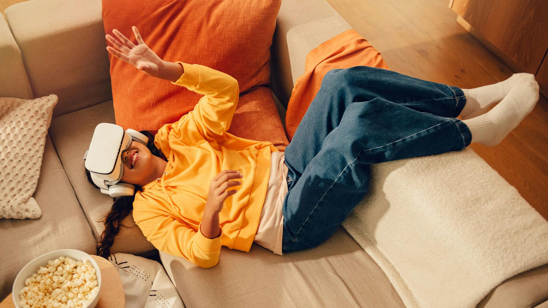 En tjej ligger i soffan med VR-glasögon och sträcker ut armen framför sig.
